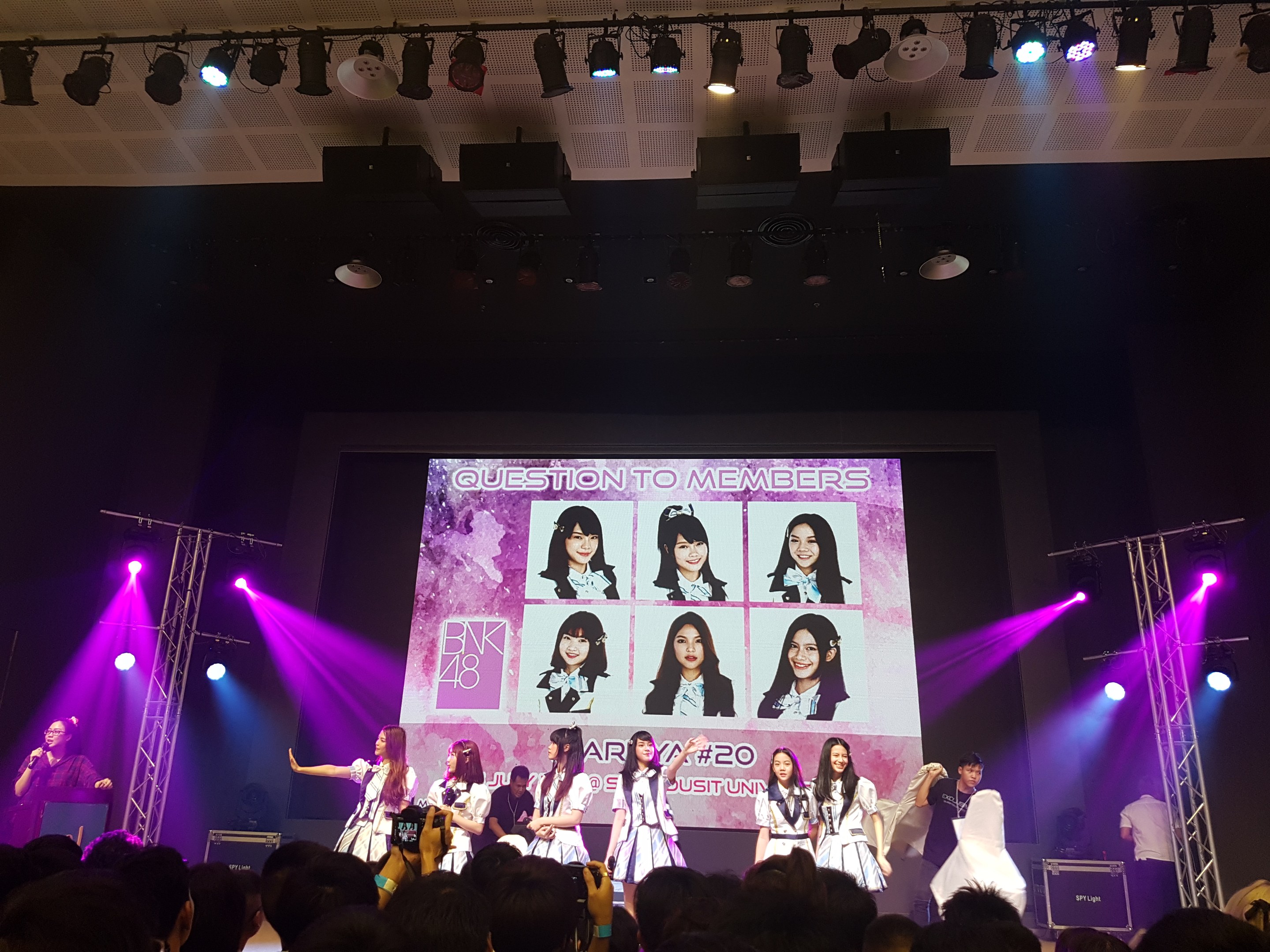 Eine Gruppe von Menschen auf einer hell erleuchteten Bühne mit Trägern, wobei einer ein Mikrofon hält und ein anderer ein Instrument spielt, während unten eine Menge Fotos macht; ein großer Bildschirm im Hintergrund zeigt Bilder der K-Pop-Gruppe BK48.