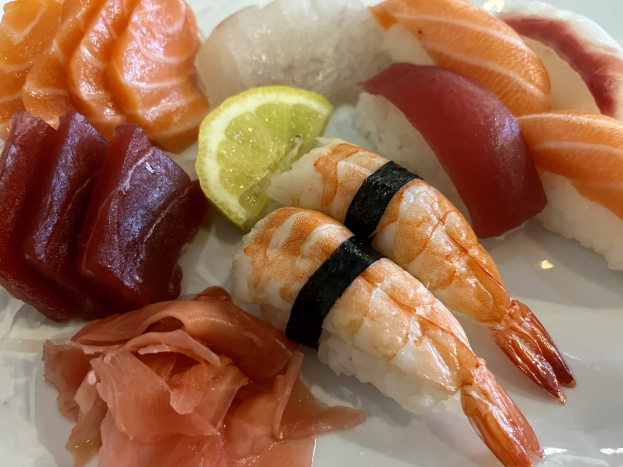 Ein weißer Teller mit verschiedenen Sushi-Stücken, darunter Lachs, Thunfisch und Garnelen, begleitet von einer Zitronenscheibe.