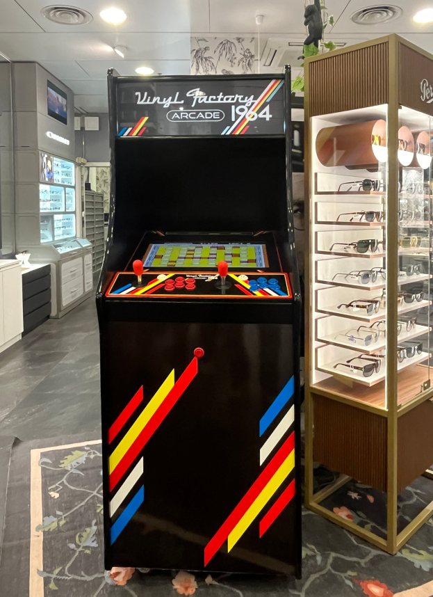 Ein Geschäft mit einem zentralen Vintage-Arcade-Automaten, einem Glastisch mit einer Glasvitrine auf der rechten Seite und einer Retro-Beleuchtung mit Schränken und einem Fernseher im Hintergrund.