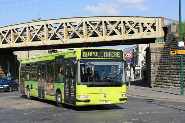 Grüner Bus fährt unter einer Brücke auf einer Stadtstraße mit Passagieren, anderen Fahrzeugen, einem Pfahl mit Schildern, einer Wand, Gebäuden, Bäumen und einem bewölkten Himmel im Hintergrund.