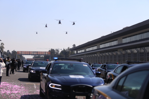 Eine Gruppe von Polizeiwagen vor einem Gebäude geparkt, mit Menschen auf der linken Seite der Straße, Bäumen, Polen, einer Brücke und einem klaren blauen Himmel im Hintergrund und vier Hubschraubern, die oben im Bild fliegen.