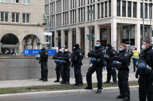 Polizisten in schwarzen Uniformen und Masken stehen vor einem Berliner Gebäude mit Glasfenstern, Säulen und umgebender Infrastruktur.