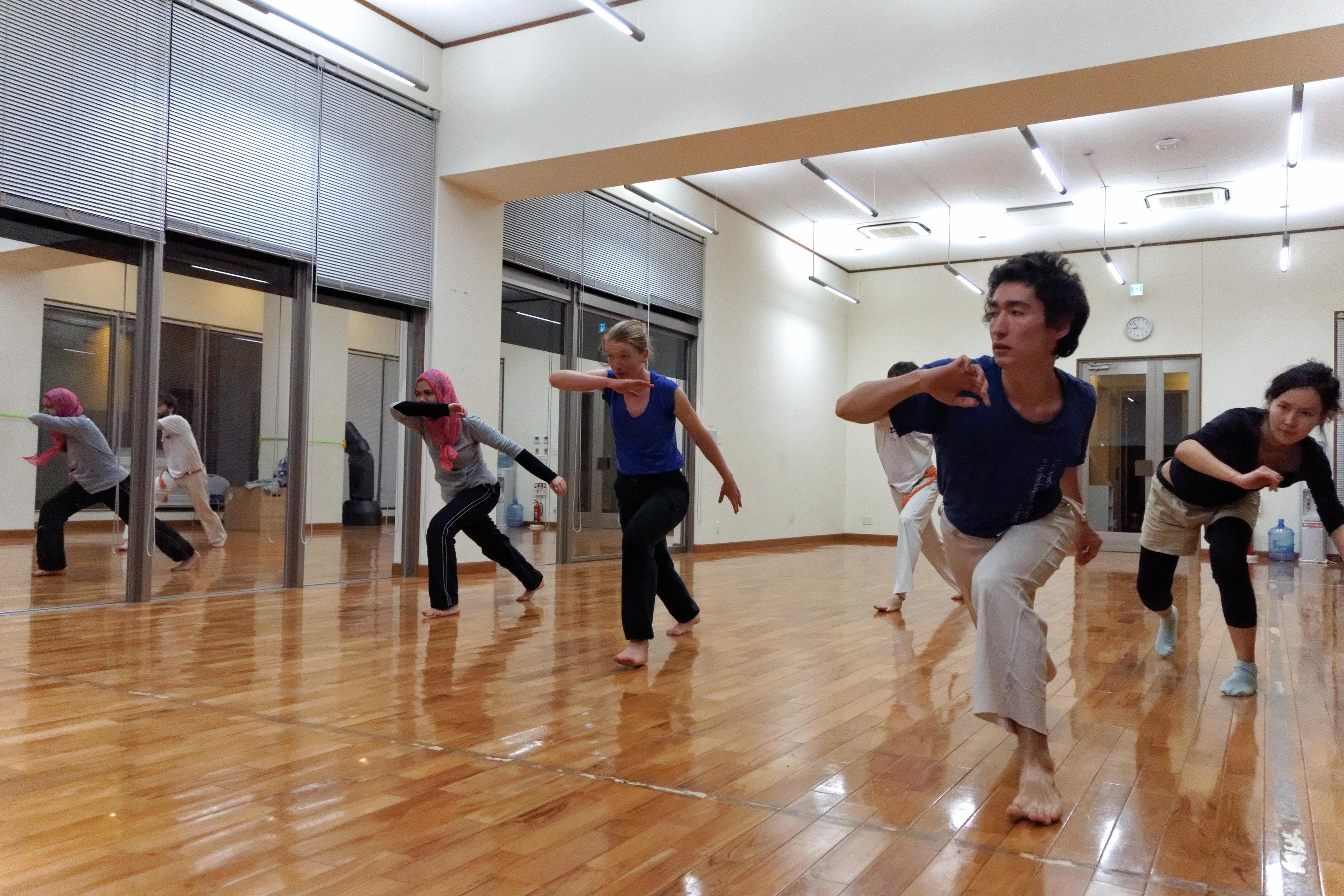 Eine Gruppe von Menschen in einem Tanzkurs mit einem Mann und einer Frau in der Mitte, umgeben von Spiegelwänden und Deckenlampen, mit einer Uhr im Hintergrund.