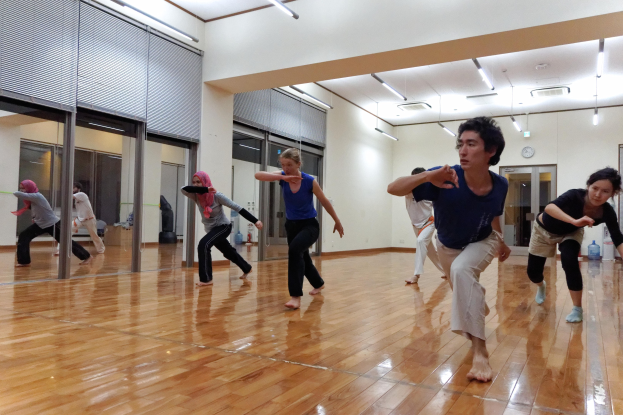 Eine Gruppe von Menschen in einem Tanzkurs mit einem Mann und einer Frau in der Mitte, umgeben von Spiegelwänden und Deckenlampen, mit einer Uhr im Hintergrund.