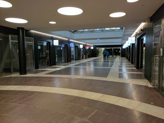 Langer Gang mit Menschen, Deckenlampen, Säulen, Glastüren und Informationsschildern, ähnlich einem belebten Rathaus.