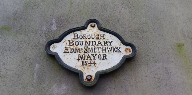 Metallplatte an einer Gebäudeseite mit schwarzem Hintergrund und weißer fettgedruckter Schrift, die "Gemeindegrenze Edm Smithwick Bürgermeister" liest.