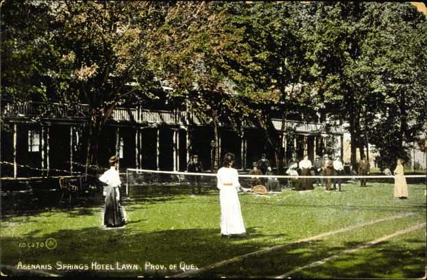 Schwarz-weißes Foto einer Gruppe von Menschen, die Tennis auf dem Rasen vor dem Abenakis Springs Hotel Lawn in Provo, Quebec spielen, mit einem Netz, sitzenden Zuschauern, Bäumen und einem Gebäude im Hintergrund.