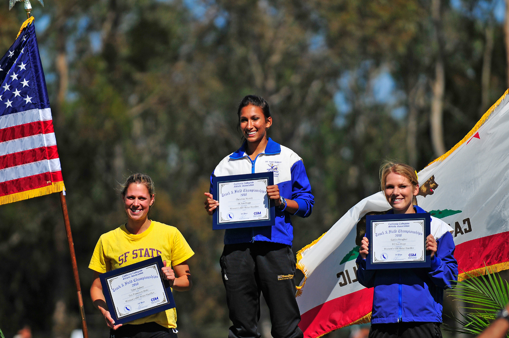 Drei Frauen mit Zertifikaten und Flaggen im Hintergrund.