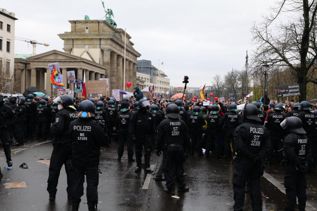 Eine große Gruppe von Polizisten steht vor einer Menge mit Schildern, Luftballons und Helmen, vor Gebäuden, Bäumen, einem Kran und einem bewölkten Himmel.