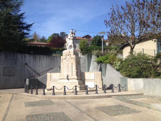 Bronzestatue eines Mannes auf einem Sockel, umgeben von einem Metallzaun, Bäumen und Pflanzen in einem Park, mit Gebäuden und einem bewölkten Himmel im Hintergrund, die als Denkmal für die Opfer der Pariser Anschläge gilt.