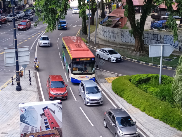 Eine belebte Stadtstraße mit Autos, Bussen, Motorrädern und verschiedenen städtischen Elementen wie Pfosten, Gebäuden und Grünflächen.