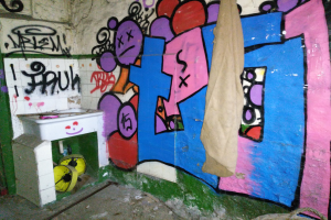 Badezimmer mit beschmierten Wänden, einem in der Ecke mit einem Tuch bedeckten Waschbecken und verstreuten Gegenständen auf dem Boden.