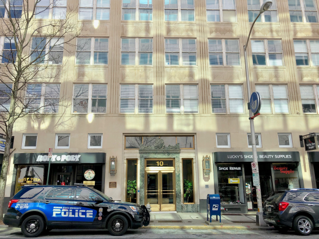 Ein Polizeiwagen vor einem Gläsgebäude geparkt, umgeben von Bäumen, Laternenmästen und Schildern, mit einem Müllkönner im Vordergrund.