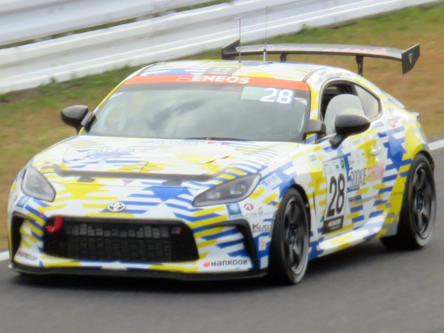 Ein Toyota Supra GT3-Rennwagen auf einer Rennstrecke mit Sponsor-Text und -Nummern, einer Begrenzung und Gras im Hintergrund.