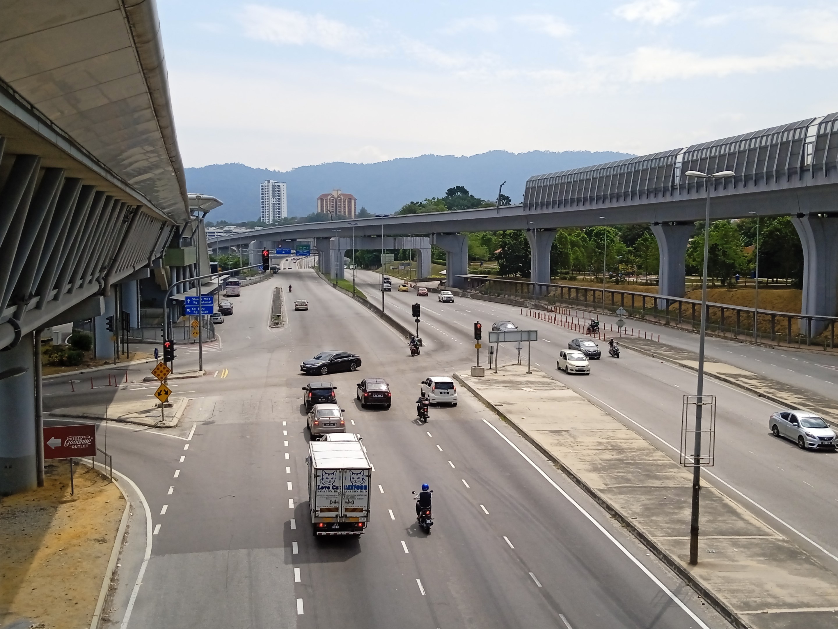 Eine vielbefahrene Autobahn mit mehreren Fahrzeugen, neben einer Brücke, mit Straßenlaternen, Verkehrsampeln, Schildern, Bäumen, Gebäuden, Hügeln und einem bewölkten Himmel.