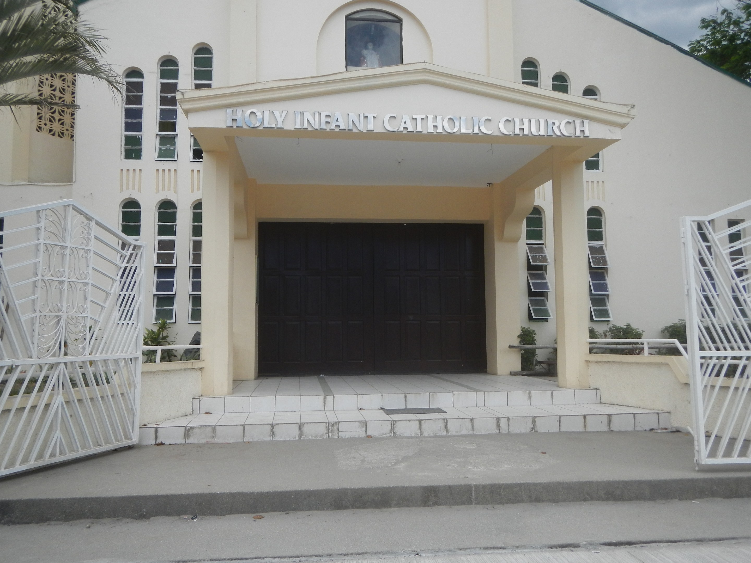 Übersicht des Heiligen-Kind-Katholischen-Kirchengebäudes mit Treppe, Geländern, umgebender Vegetation und einem bewölktem Himmel.