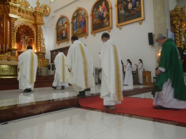 Eine Gruppe von Priestern geht einen roten Teppich in einer Kirche entlang, mit Menschen, die auf dem Boden und an den Wänden mit Foto-Rahmen im Hintergrund stehen.