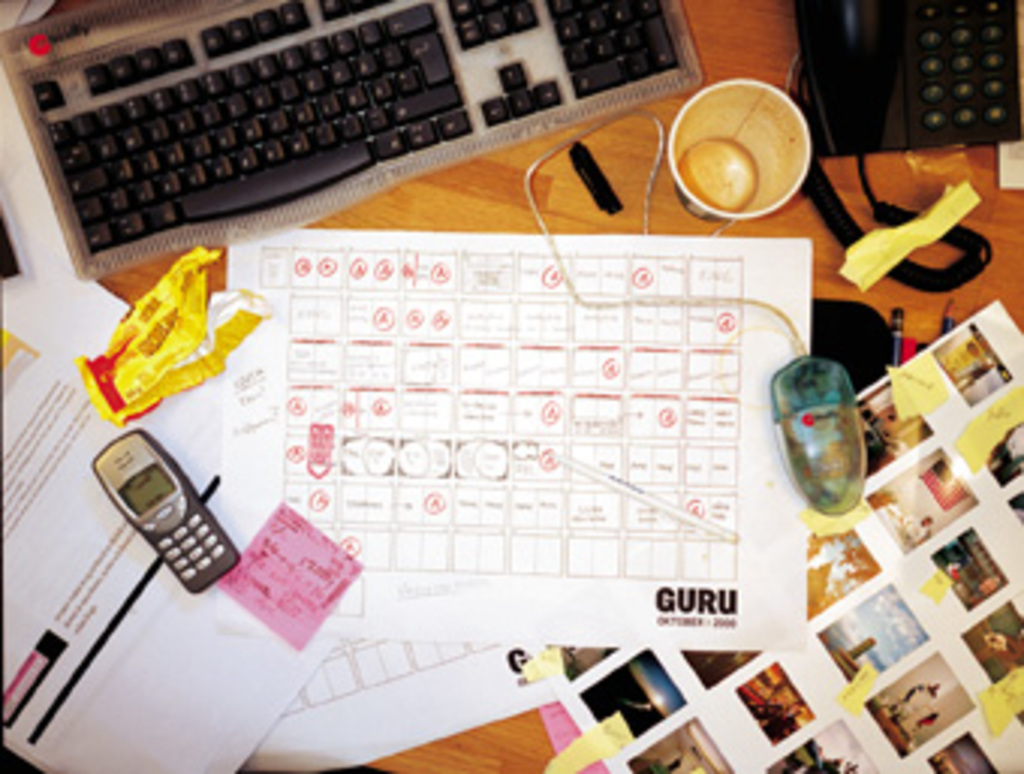 Ein chaotischer Schreibtisch mit Papieren, einem Computer, einem Telefon, Tassen und einem Handy.