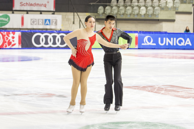 Zwei Eiskunstläufer, ein Mann und eine Frau, gleiten Hand in Hand über eine Eisfläche mit einer Menge und Absperrungen im Hintergrund.