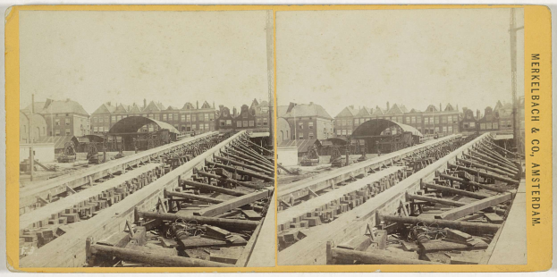 Schwarz-weißes Foto einer Zugstrecke mit Gebäuden im Hintergrund, Text 'Merkelbach & Co, Co. Amsterdam' auf der rechten Seite und verstreute hölzerne Gegenstände im Vordergrund.