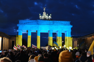 Eine Menschenmenge steht vor dem Brandenburger Tor in Berlin, Deutschland, mit Fahnen und Plakaten in den Händen, mit einem Banner auf der rechten Seite und dem Tor mit Statuen und Säulen geschmückt unter einem bewölkten Himmel.