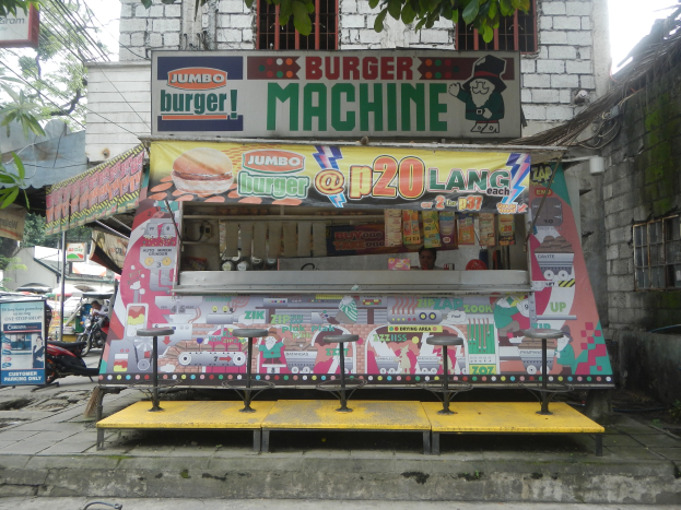 Ein leuchtend gelber und roter Foodtruck in Form einer Jumbo-Burger-Maschine parkt auf einer Stadtstraße, umgeben von Gebäuden, Bäumen und Menschen.