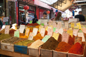 Ein belebter Marktplatz mit zahlreichen Gewürzkisten, jede mit einem Preisetikett versehen, und Menschen im Hintergrund unter Deckenbeleuchtung, mit einem Stand auf der linken Seite, der verschiedene Artikel anbietet.