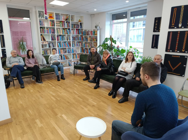 Eine Gruppe von Menschen sitzt um einen Tisch in einem Raum mit Bücherregalen, Pflanzen und einem Fenster, die während eines Workshops zu Gesundheit und Wohlbefinden in Berlin diskutieren.