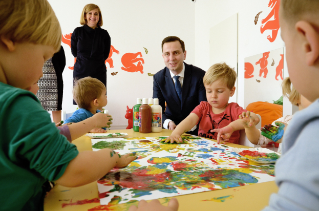 Gruppe von Kindern bei einer Malaktion um einen Tisch mit bunten Handabdrücken, Papieren und Flaschen, während eine Frau im Hintergrund mit Kunstwerken an der Wand lächelt.