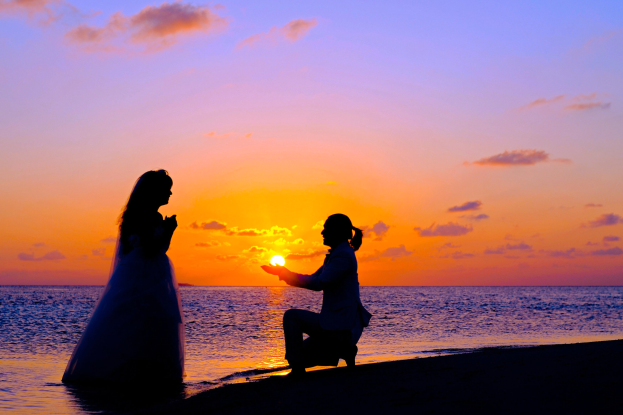 Braut in weißem Kleid und Bräutigam, der auf dem Strand in der Hocke ist, bei Sonnenuntergang, Sonne tief am Himmel hinter ihnen, Himmel in Orangen, Pinks und Purpur, tiefes blaues Wasser.