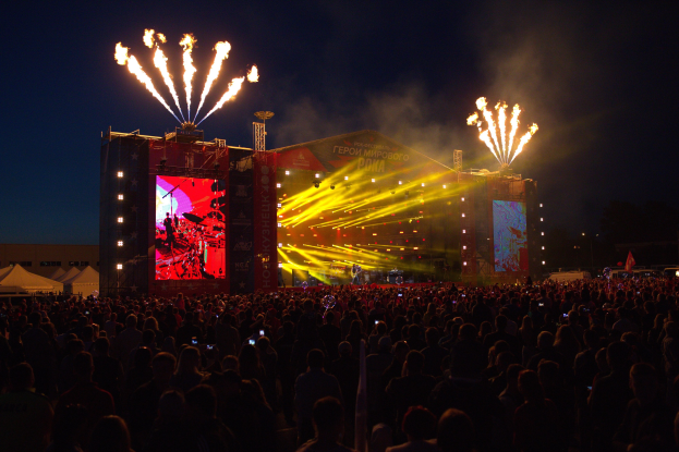 Eine große Menschenmenge auf einem Musikfestival schaut auf einer hell erleuchteten Bühne Performern zu, mit Zelten links und Feuerwerk im dunklen Himmel dahinter.