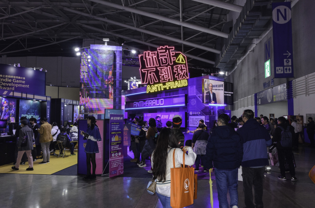 Eine große Gruppe von Menschen steht vor einem "Indie Game Development Award 2019"-Stand an einer Messe, wobei einige Taschen halten, während Bretter, Bildschirme und Lichter im Hintergrund zu sehen sind.