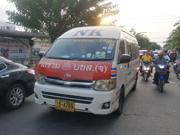 Ein weißer Lieferwagen fährt eine Straße mit parkenden Autos und Motorrädern entlang, mit Menschen in Helmen, Bäumen, Gebäuden, Texttafeln, Polen, Drähten und einem klaren blauen Himmel im Hintergrund.