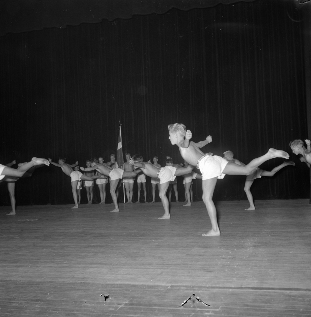 Schwarzes Foto einer Gruppe, die auf einer Bühne mit Vorhang und Flaggenhintergrund Gymnastik macht.