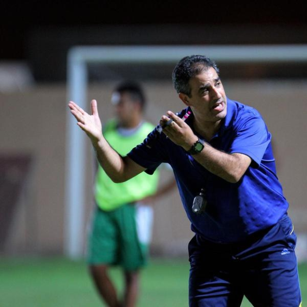 Ein Fußballtrainer in einem blauen T-Shirt und Hose steht auf einem Feld und hält ein Mikrofon in der Hand, während er auf Spieler zeigt, mit einem Torpfosten und einer Person in einem grünen Kleid im Hintergrund.