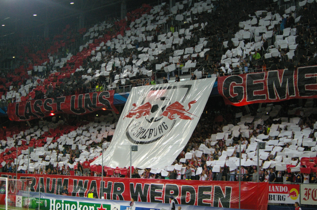 Ein Fußballstadion mit sitzenden und stehenden Zuschauern, das Banner, ein Torpfosten und Flutlicht zeigt.