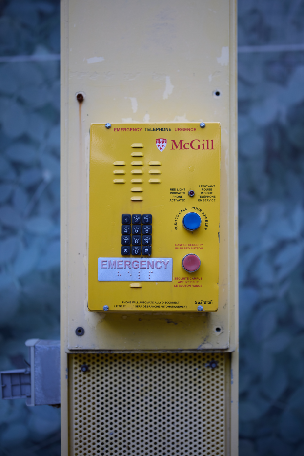 Ein gelber Notrufhörer an einer Gebäudeseite angebracht, mit Tasten und Text für Notrufe, vor einer Wand.