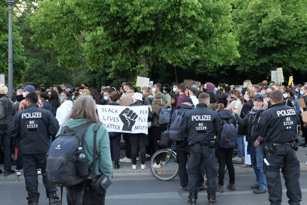 Eine große Gruppe von Menschen nimmt an einer Black Lives Matter Demonstration in Berlin teil, einige halten Schilder und tragen Mützen und Taschen, mit einem Fahrrad im Vordergrund und Bäumen und einem Pfahl im Hintergrund.