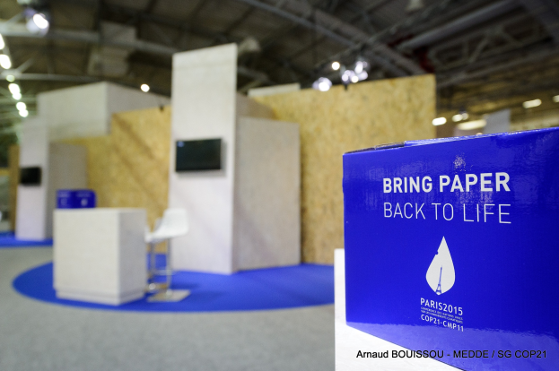 Messestände mit blauer und weißer "Bring Paper Back to Life"-Aufschrift auf der rechten Seite, mit unscharfen Stühlen, Tischen, einem wandmontierten Fernseher und Deckenlampen im Hintergrund.