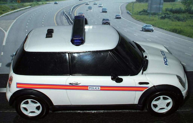 Weißer Polizeiwagen mit "Polizei"-Aufschrift auf einer Straße außerhalb einer Stadt, umgeben von fahrendem Verkehr, Gras und Bäumen auf beiden Seiten.