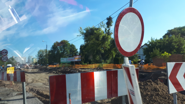 Baustelle mit einem roten und weißen Schild am Straßenrand, Bäume, Laternen, ein Verkehrszeichen im Vordergrund, Gebäude im Hintergrund und einen klaren blauen Himmel.