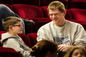 Ein Mann und zwei Kinder mit Brille und strahlendem Lächeln sitzen in einem Kinosaal mit zusätzlichen Stühlen im Hintergrund.