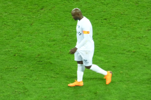 Ein Fussballspieler in einem wei├čen T-Shirt, wei├čen Shorts und orangenen Schuhen ├╝berquert ein saftig gr├╝nes Feld.