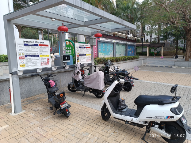 Elektrische Scooter vor einem Bushaltestellen mit nahen Informationsschildern umgeben von Bäumen, Gebäuden und einem klaren blauen Himmel.