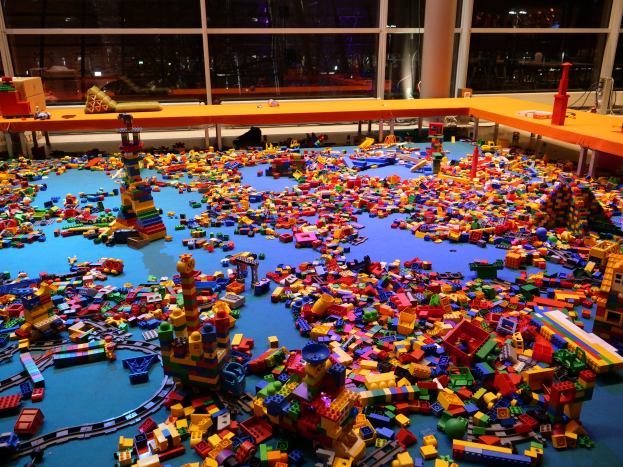 Ein großer Raum mit farbigen Lego-Steinen auf dem Boden, umgeben von Tischen und Säulen, mit Glasfenstern im Hintergrund, die Menschen und Lichter zeigen.