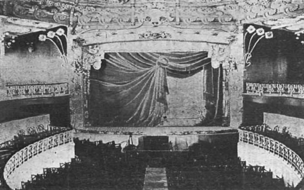 Schwarzes und weißes Foto der Theater Royal-Bühne in London, das eine Bühne mit Vorhang, Geländer und ornamentierte Wandgestaltung zeigt, mit ein paar sitzenden Menschen und Text am unteren Rand.