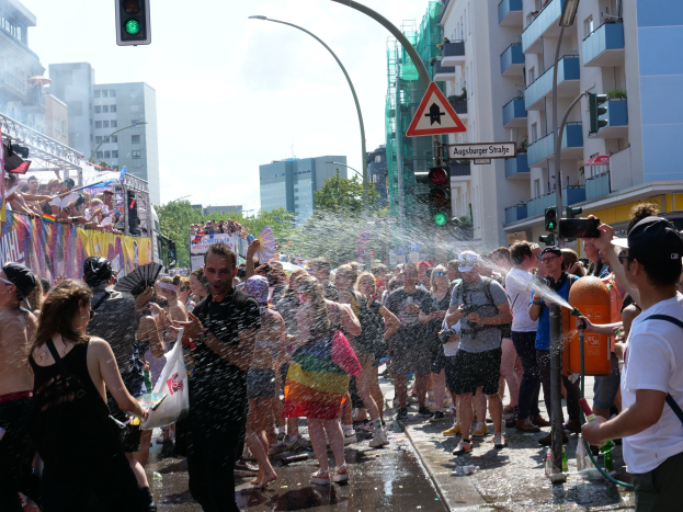 Menschen auf einer Straße während eines Christopher Street Day-Umzugs, einige sprühen Wasser und halten Gegenstände, mit einem Banner, Gebäuden, Bäumen und Ampeln im Hintergrund.