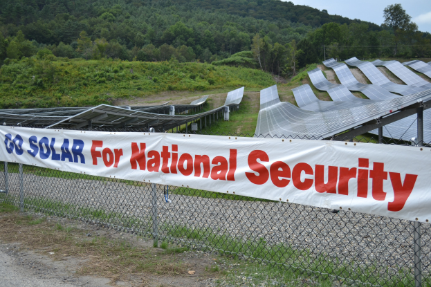 Ein Zaun mit einer 'Geht Solar für die nationale Sicherheit' -Plakette vor einem Feld aus Solarpanelen, Bäumen, Pflanzen, Gras und einem klaren blauen Himmel.