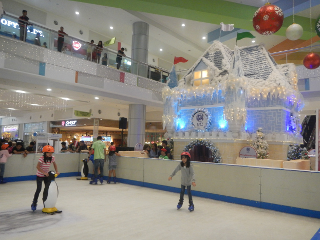 Menschen auf einer Indoor-Eislaufbahn in einem Einkaufszentrum umgeben von festlichen Dekorationen, darunter eine hauseigene Struktur mit Lichtern und einer Menge, die von Geländern und Säulen aus zusieht.