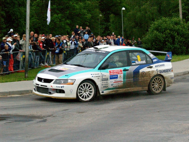 Ein Rallyefahrzeug mit Text und Logos fährt an einer Menge vorbei, die Kameras hält, mit Geländern, Fahnenmasten und Bäumen im Hintergrund.